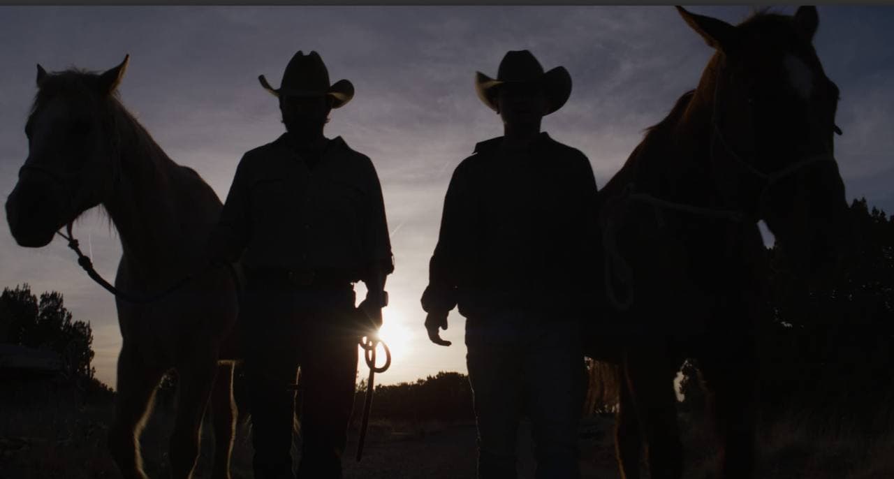 Two men in cowboy hats lead horses toward the camera, silhouetted against the setting sun.