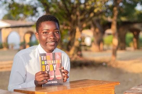 A student in Chitipa, Malawi, smiling and holding a book at an outdoor desk.